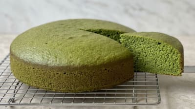 Gâteau moelleux au matcha entier posé sur une grille de refroidissement métallique, avec une part découpée révélant la mie d'un vert jade homogène, fond de marbre blanc