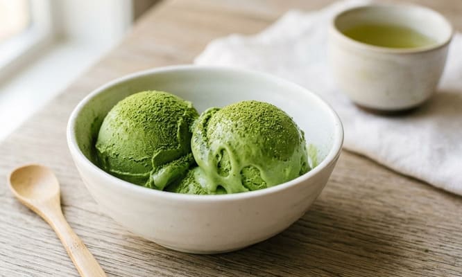 Trois boules de glace au matcha maison saupoudrées de poudre de matcha dans un bol blanc en céramique, cuillère en bambou et tasse de matcha en arrière-plan flou