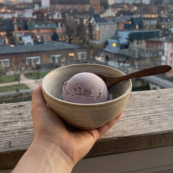 Main tenant un bol en céramique mouchetée contenant une boule de glace à l'ube violette avec cuillère en bois, vue panoramique floue sur la ville en arrière-plan au coucher du jour