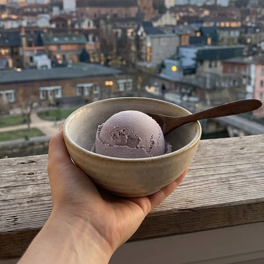 Main tenant un bol en céramique mouchetée contenant une boule de glace à l'ube violette avec cuillère en bois, vue panoramique floue sur la ville en arrière-plan au coucher du jour