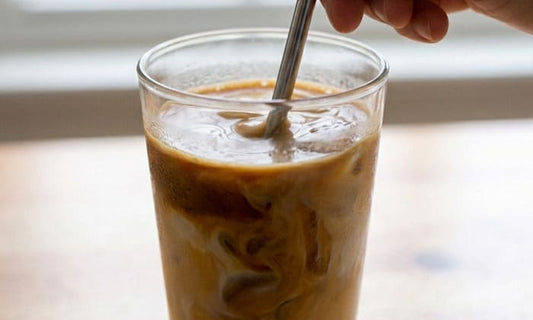 Gros plan sur un verre de iced hojicha latte ambré avec un effet marbré lait et hojicha, une main mélangeant avec une paille en inox devant une fenêtre lumineuse