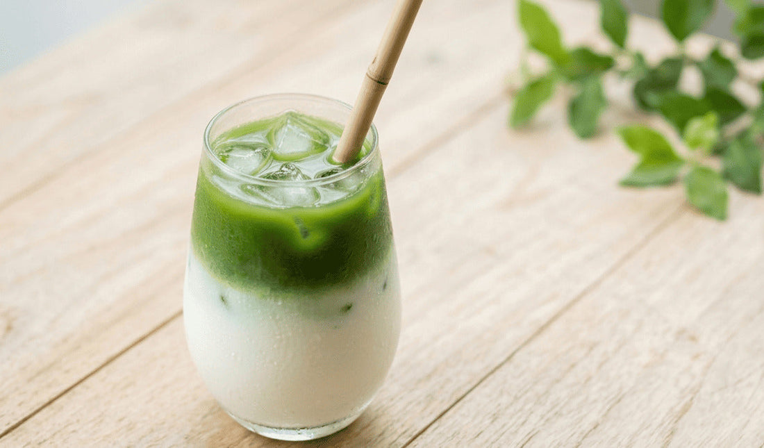 Iced pandan latte classique dans un verre arrondi transparent avec dégradé vert vif sur lait blanc et glaçons visibles, paille en bambou, branche de menthe fraîche en arrière-plan flou sur surface en bois