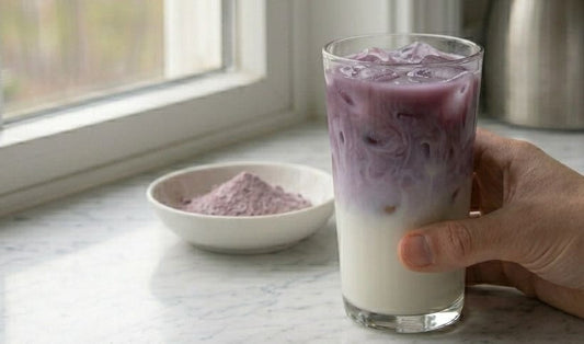 Main tenant un grand verre d'iced ube latte avec dégradé naturel violet sur lait blanc et glaçons, bol de poudre d'ube violette pâle posé en arrière-plan sur marbre blanc, lumière naturelle de fenêtre